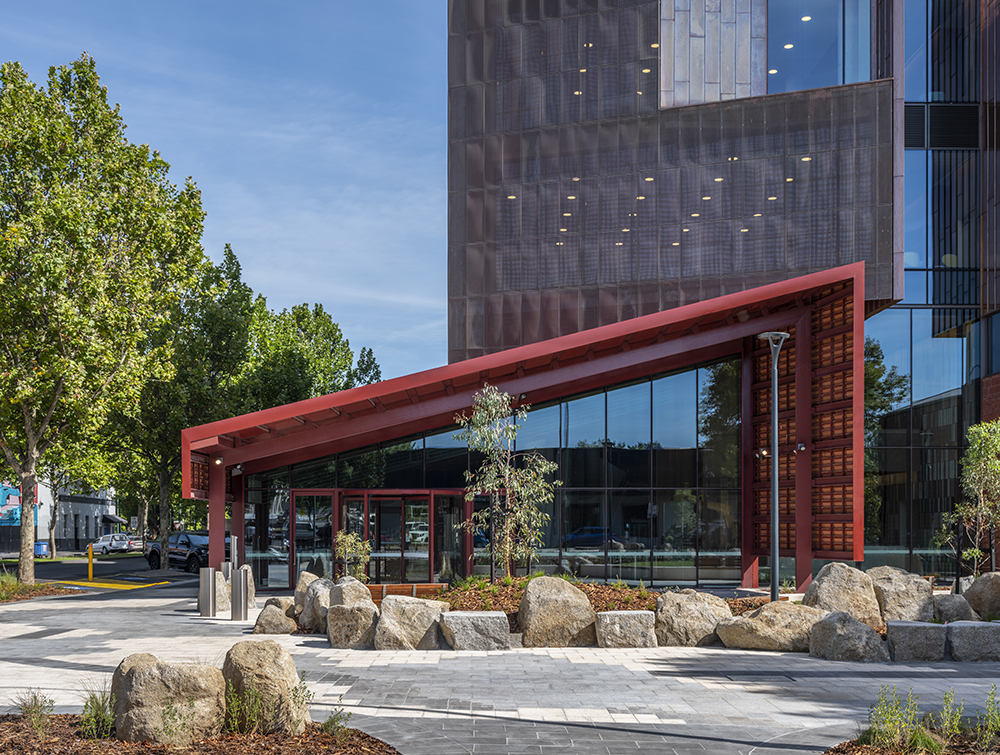 A paved and landscaped area looks towards a vaulted awning sitting over glass doors